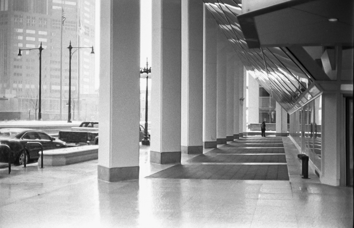 A lone figure beneath a repeating row of pillars, with winter light falling across the sidewalk.